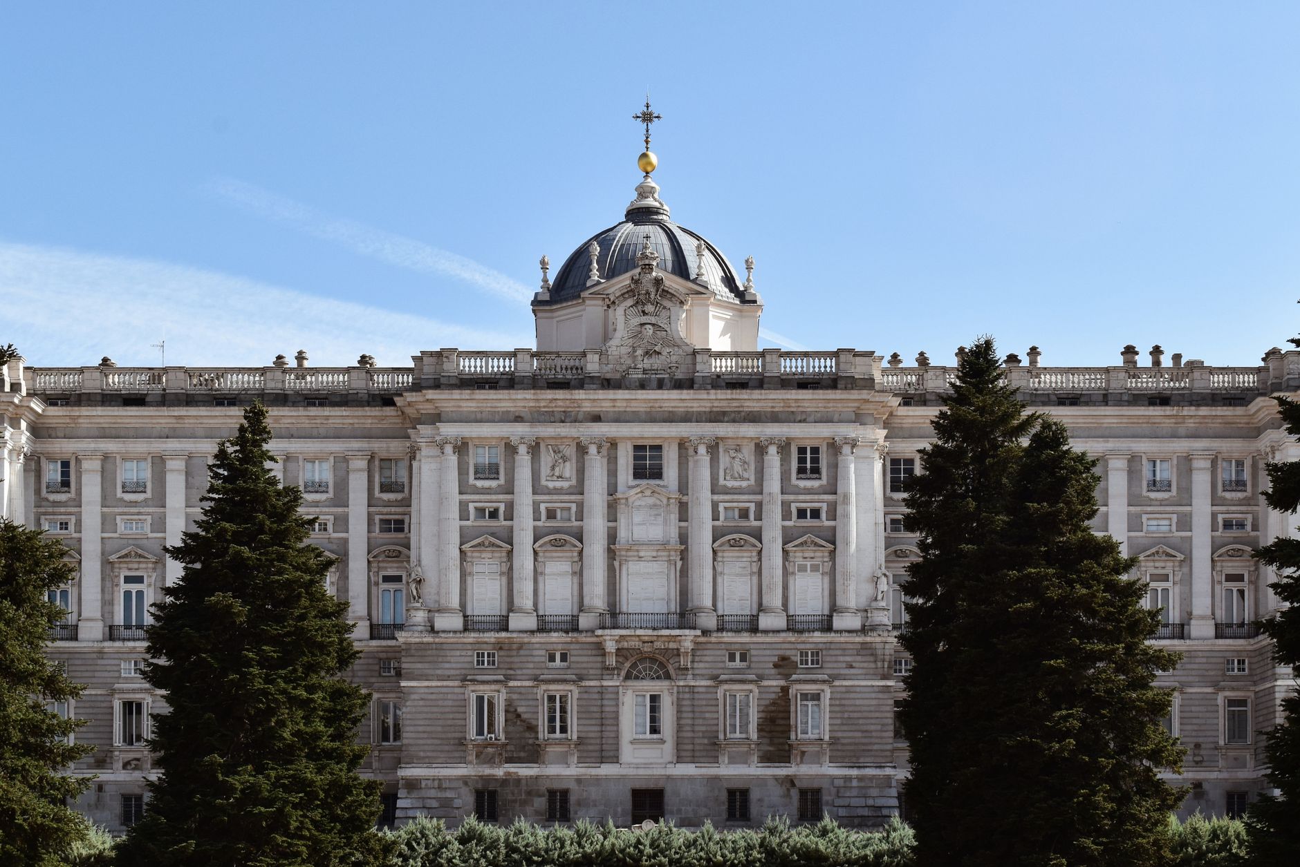 The grand facade of the Royal Palace of Madrid under a clear blue sky, framed by lush trees.